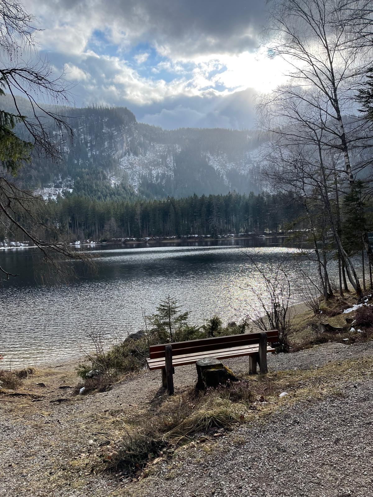 Holzbank am winterlichen Altausseer See, dahinter Sonnenstrahlen über den verschneiten Bergen