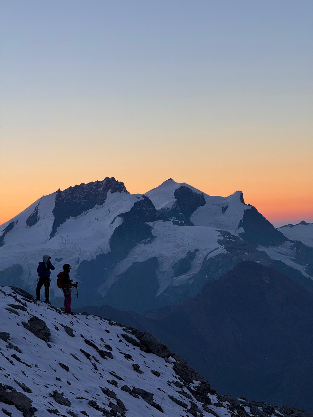 Zwei Winterwanderer auf einem Schneegrat vor Gipfeln im Morgenrot